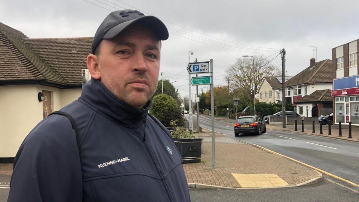 Clive Naylor wears a dark blue jacket and a charcoal coloured cap. He is standing on the pavement next to a main road opposite a row of shops.