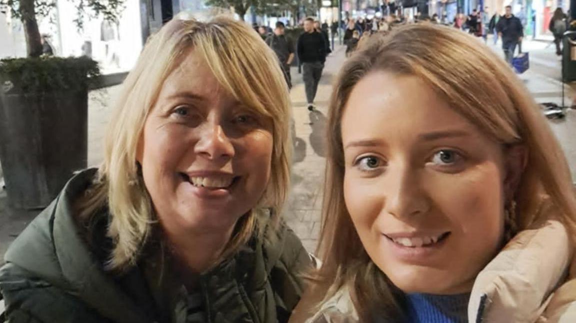 Sharon McDowell on the left has shoulder length blonde hair and is wearing a green coat. Claire McDowell on the right has long light ginger hair and is wearing a cream coat. 