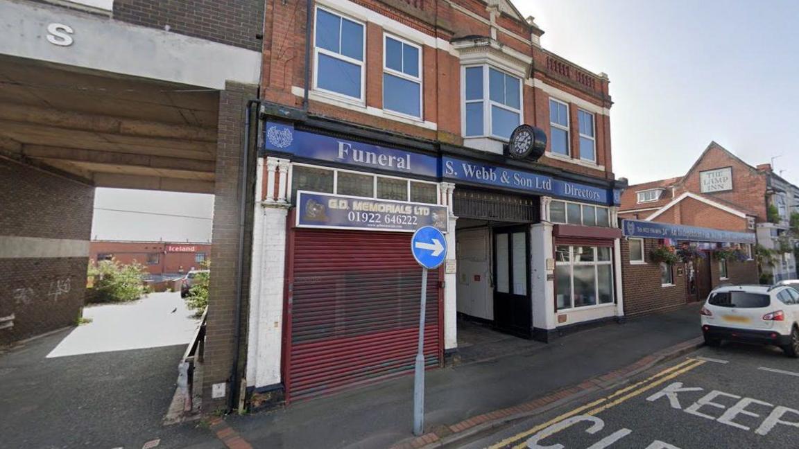 A Google street view image of the front of S. Webb & Son Funeral Directors in Wednesbury. The shop has a blue sign above the entrance with its name in white lettering. Cars are parked alongside the road to the right of the store.