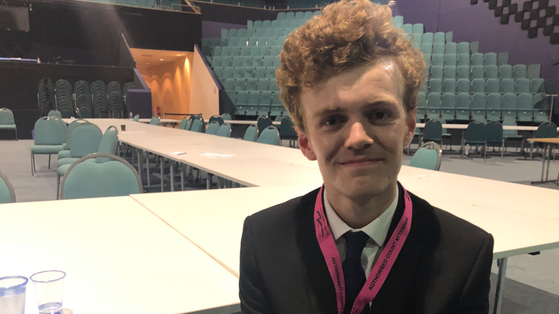 Sam Carling, in a jacket and tie, smiling in an empty hall where the election count was held.