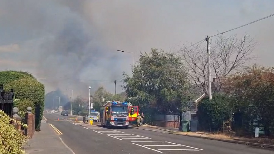 A fire engine is parked on a road with a plume of smoke seen in the distance. There are trees and houses either side of the road as well as parked vehicles further back.