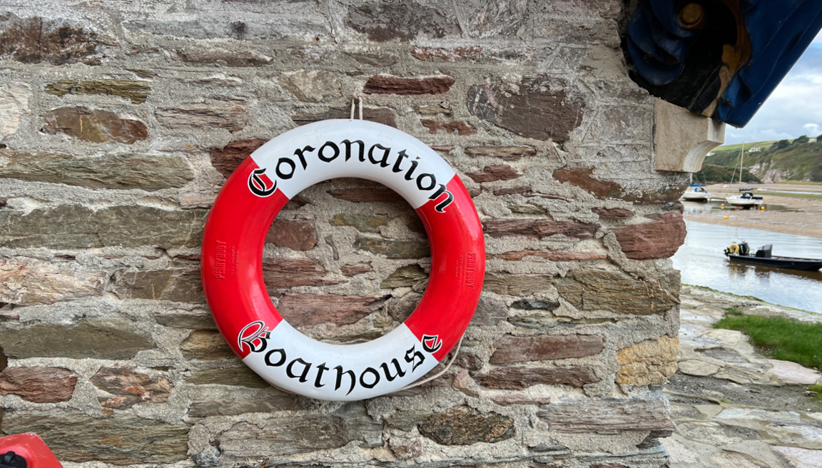 A red and white lifebelt with the wods Coronation Boathouse attached toa stone wall with a river to the right and a boat moored in the river.