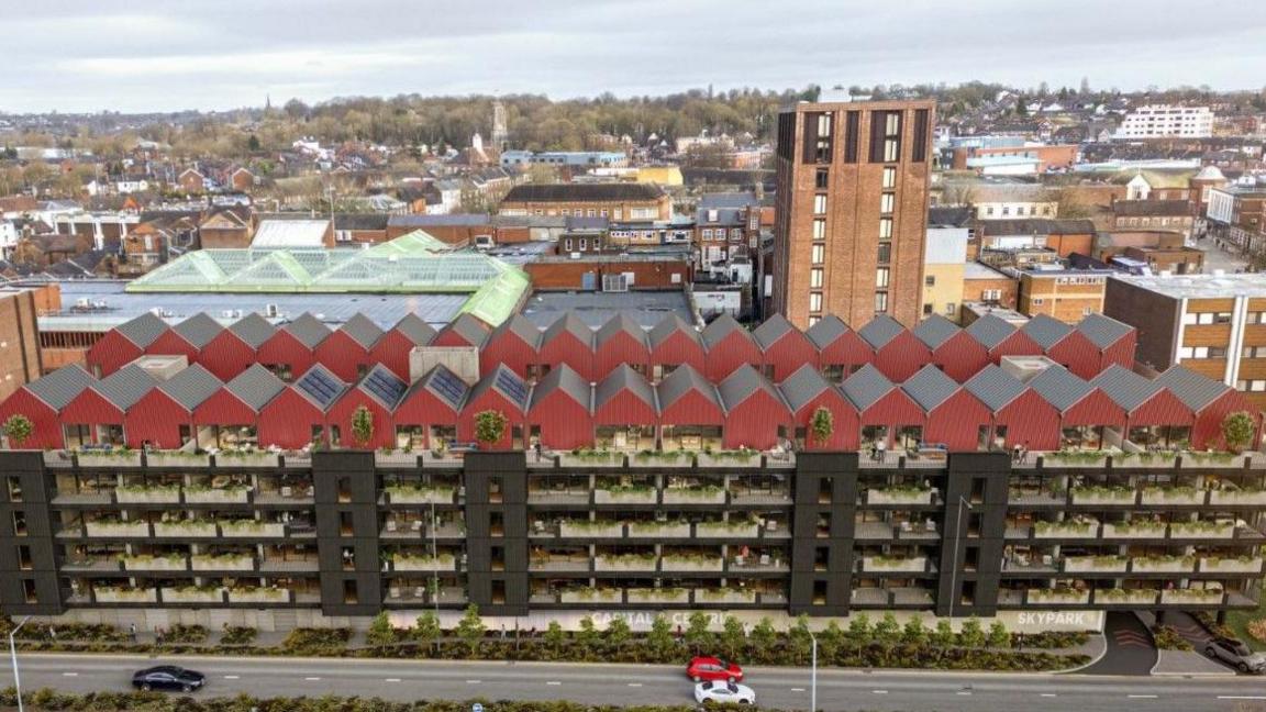 Flats over several storeys, with those on the highest level red on the outside. The road, with some cars, is at the bottom of the photo.  