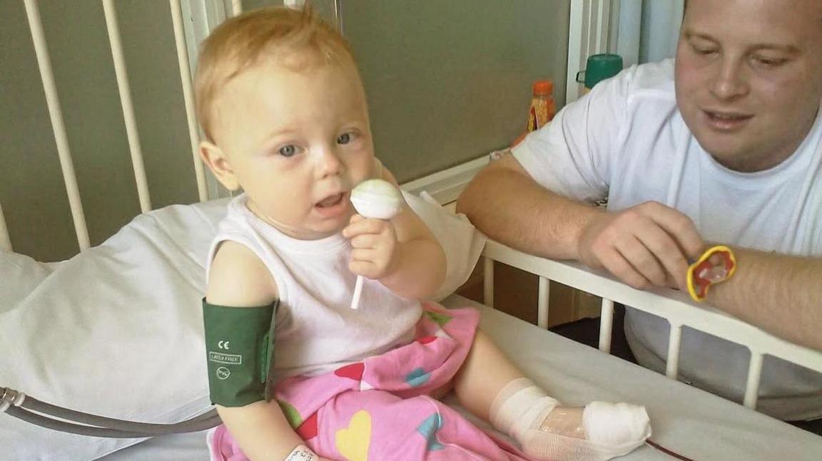 Maisie as a baby sat in a hospital bed eating a lollipop as he dad sits next to her.