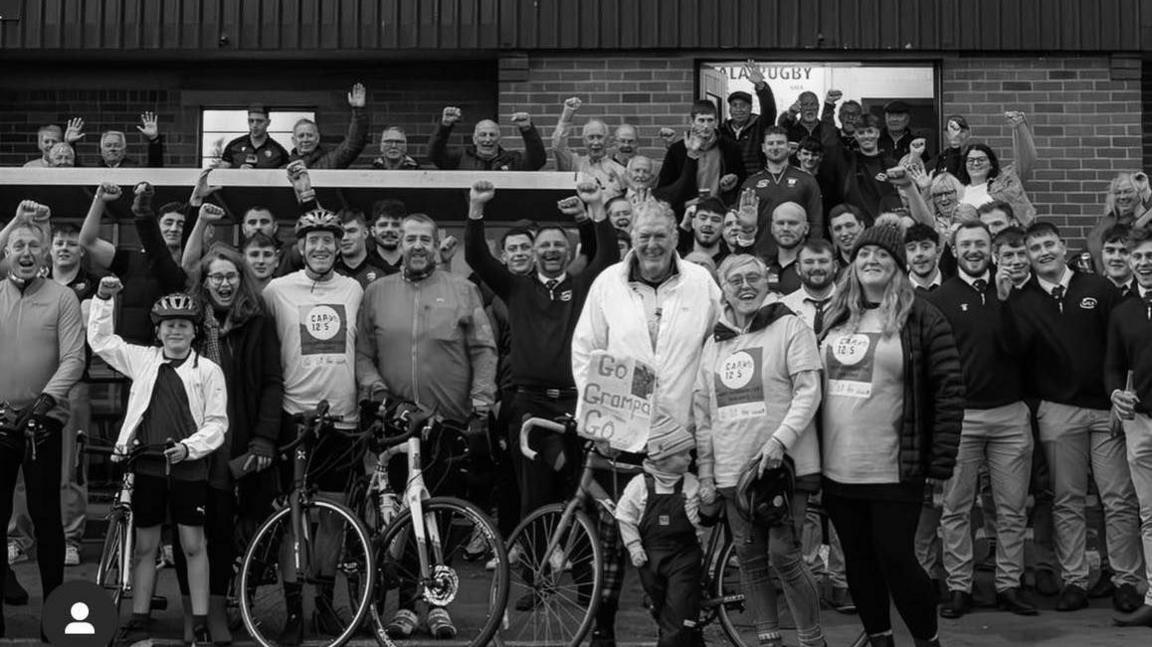 Full black and white outdoor shot of a large group of people, many on bicycles, in front of a grandstand at a sports venue. The grandstand is a multi-tiered structure with rows of light beige/cream-colored seats.