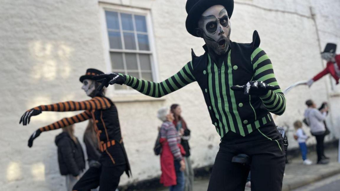 Two people dressed as skeletons walking through the street with their arms up in front of them. One is looking directly into the camera with his mouth open, he is wearing a green and black skeleton costume. The other is looking ahead of them and is wearing an orange and black skeleton costume. They both have skeleton face paint on. 