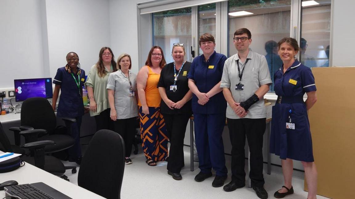 A line of people wearing various medical uniforms standing in an office with PCs, chairs and desks.