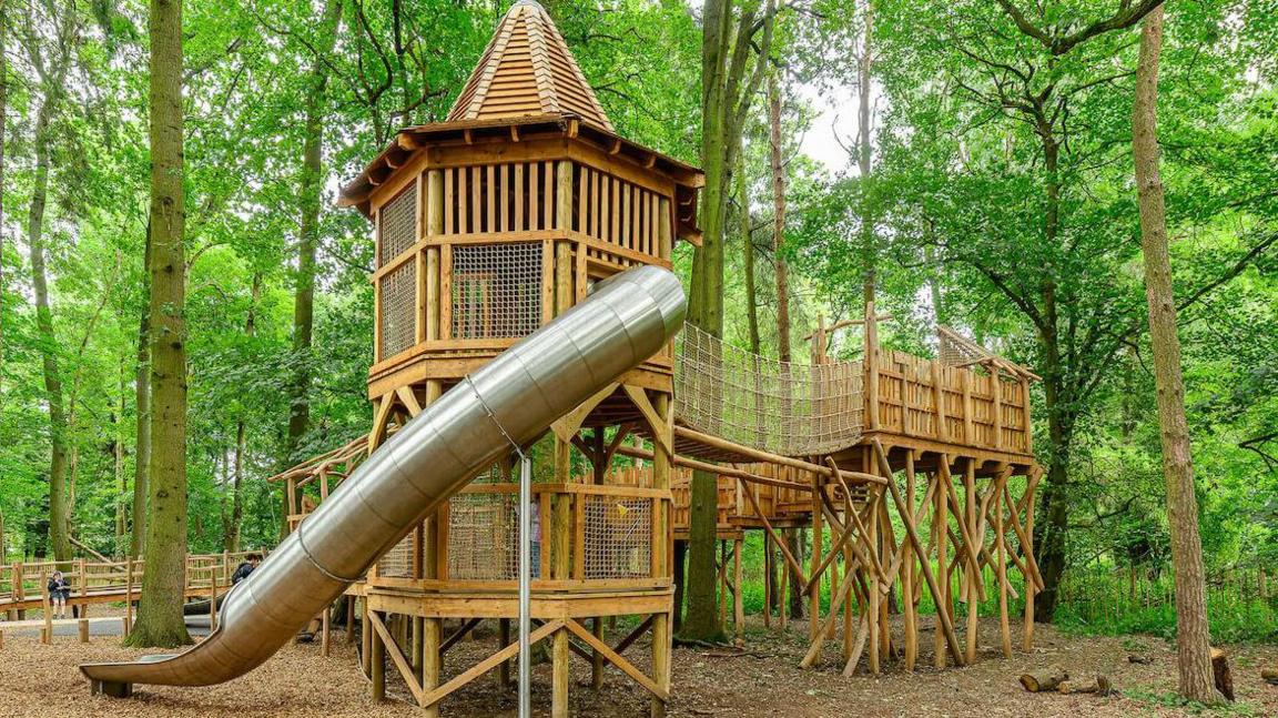 Wooden play equipment with a metal slide which is in a woodland area. It includes a climbing area and platform on wooden stilts. There are trees in the background.