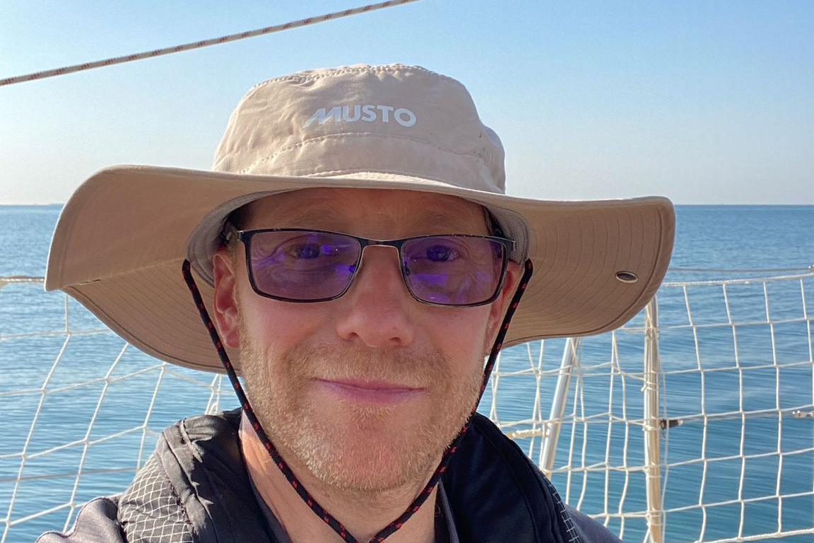 A man wearing a beige wide brim hat and sunglasses takes a selfie against the backdrop of the sea