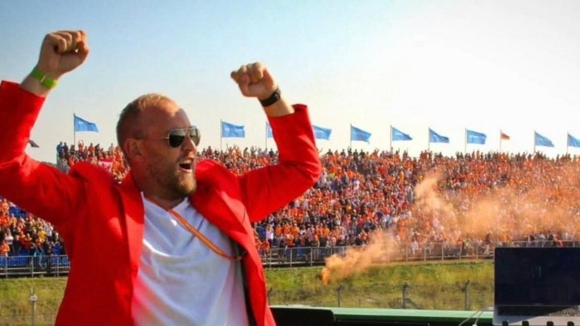 Rick Jansen DJs for the crowds at Zandvoort. Orange smoke from flares can be seen in the background, while Rick is wearing an orange jacket and is lifting his arms in the air