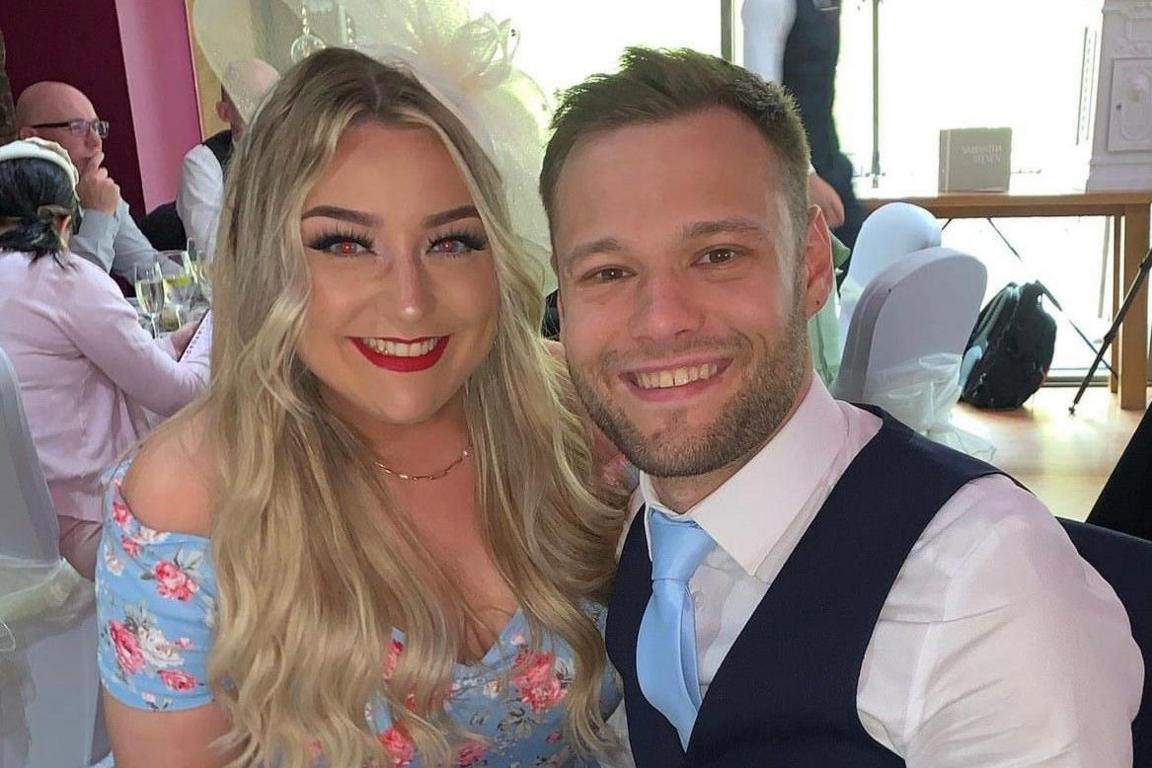 Lara is wearing a light blue dress with flowers on it. She has long blonde hair and is wearing a fascinator in her hair. She is smiling at the camera and has red lipstick on. Kieran is in a waistcoat and light blue tie. He is smiling at the camera. They are at a wedding.