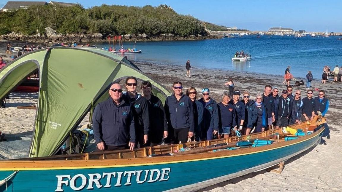 A group of people stand on a sandy beach beside a large blue wooden boat named 'FORTITUDE' with white lettering. A green tent is partially covering the boat. In the background, more people, boats, and water activities are visible in a bay or harbour, with vegetation and buildings along the distant shoreline.