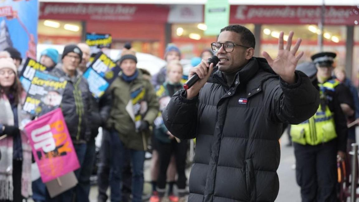 Daniel Kebede, general secretary of the National Education Union, pictured in the foreground wearing a black jacket and holding a microphone. Union members can be seen in the background.