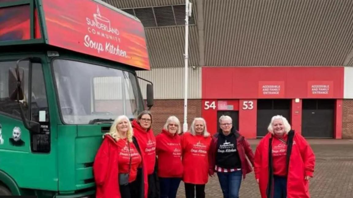 A group of six volunteers from the Sunderland Community Soup Kitchen stand in front of a green lorry which has the name of the charity on a red/orange sign above the cab. They are outside the Stadium of Light, with part of a stand and closed turnstiles behind them.
