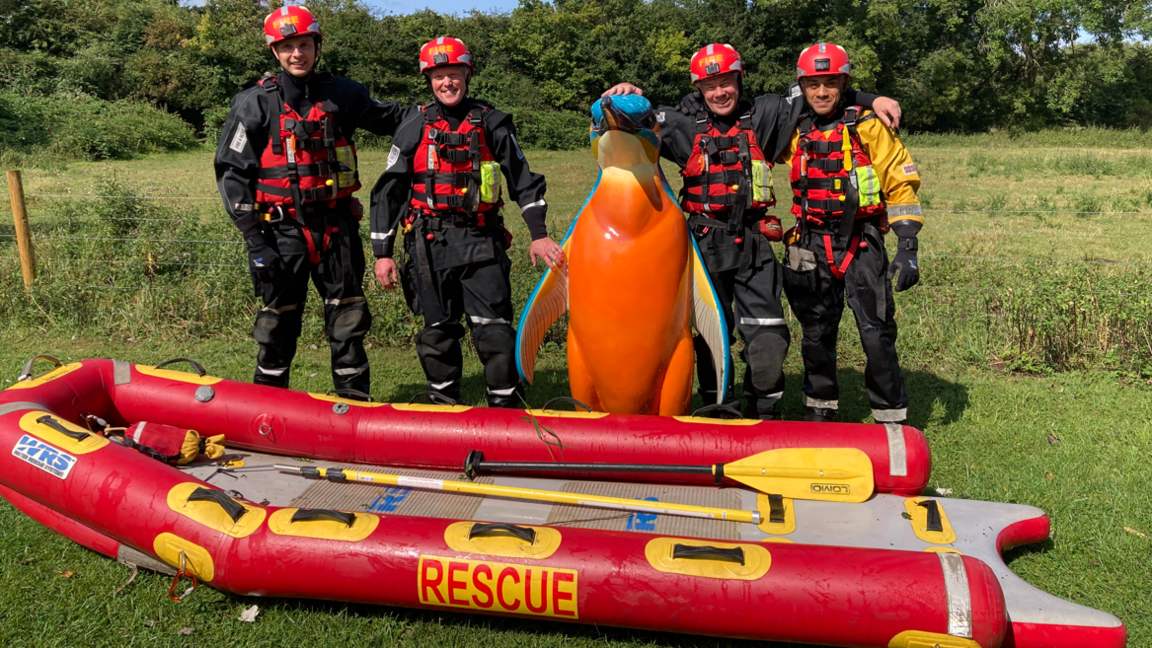 Four firefighters in black and red water rescue gear stand with a tall blue and orange penguin sculpture between them, in front of them is a red inflatable dinghy that has the word rescue written on the side