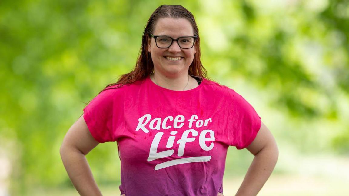 A woman wearing a pink top with "Race for Life" written on it in a white font. She is smiling and has long brown hair and black rimmed glasses.
