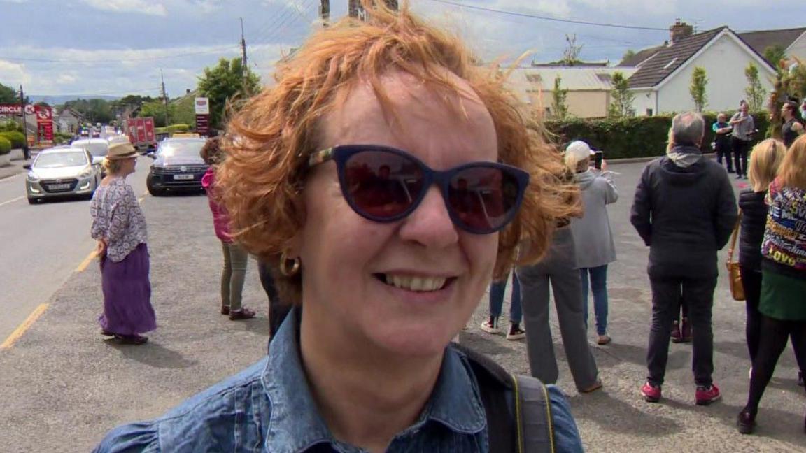 Grainne McCool, smiling, while wearing sunglasses and a denim top. She has short, wavy red hair and has the strap of a bag slung over her left shoulder. 