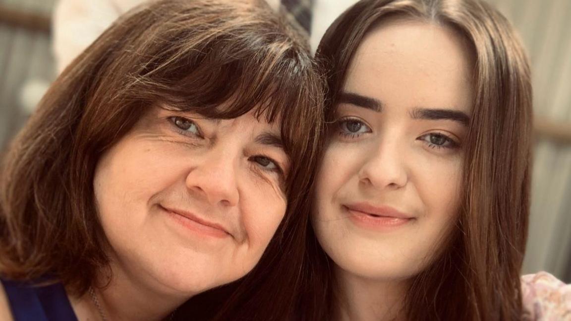 Emily and her mother are hugging and both smiling at the camera. They both have long brown hair.