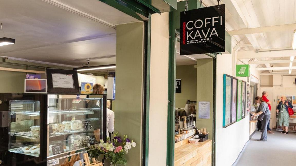 The outside of a cafe, with cake cabinets and a counter and a sign which reads "Coffi Kava".