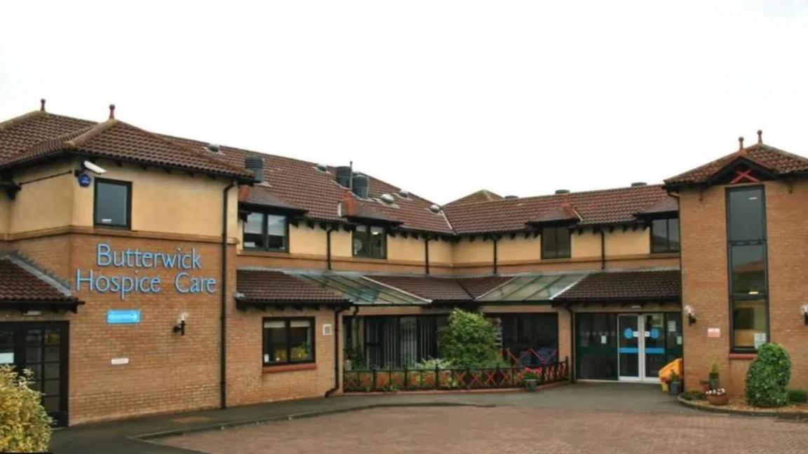 View of a two-storey building, brick on the ground floor, beige cladding on the upper, which has gable windows. It is set at a right angle, with a glassed front door to the right, and an area of shrubs in the middle. A sign identifies it as Butterwick Hospice Care. 