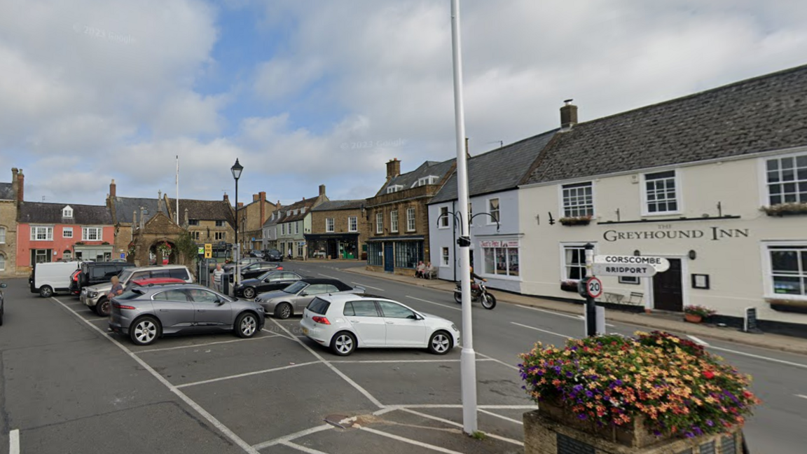 Beaminster town square with the car park in the centre. Around the edge are shops and a pub - all are old terraced buildings with pitched roofs.