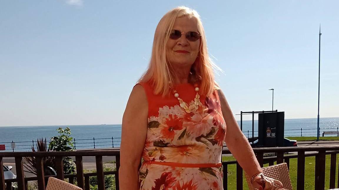A woman in a floral pink white and brown dress is posing on a balcony overlooking a seafront. She has long blonde hair, is smiling, and wearing dark circular sunglasses.