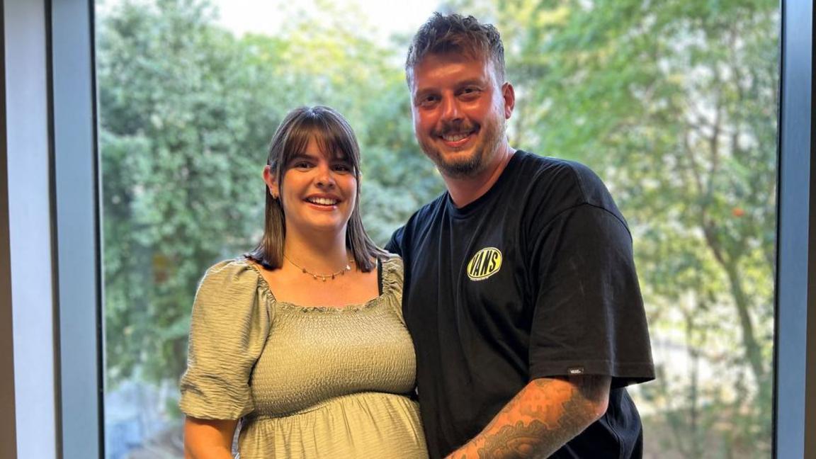 A pregnant woman in a light green dress with shoulder length brown hair and a fringe is smiling at the camera and stood up. A man next to her in a black vans top and with arm tattoos and short brown hair has his hand on her stomach and is also smiling at the camera. You can see a window and greenery behind them.