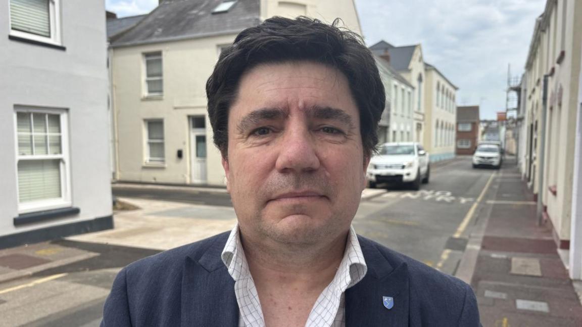 Jason Lagadu stands on a street lined with buildings on both sides. He has dark brown hair and is dressed in a dark blazer over a light-coloured, checked shirt. Along the street, there are parked cars and the sky is cloudy.
