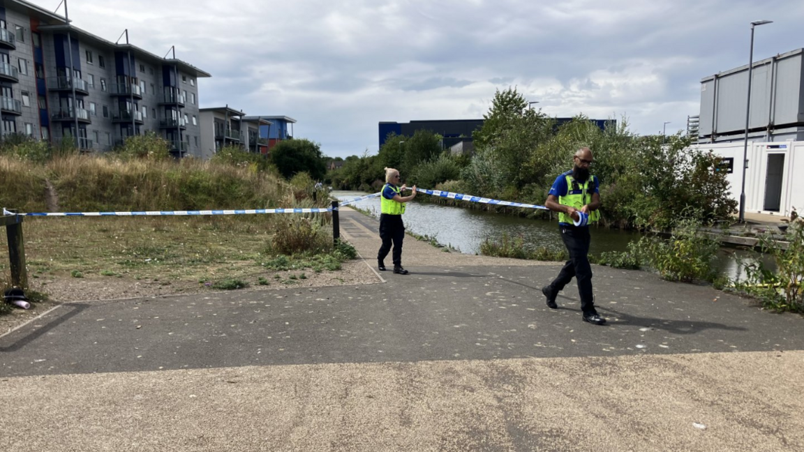 Two police officers, one male and one female, roll out police tape as they create a cordon on the canal, which has greenery on both sides and blocks of flats overlooking