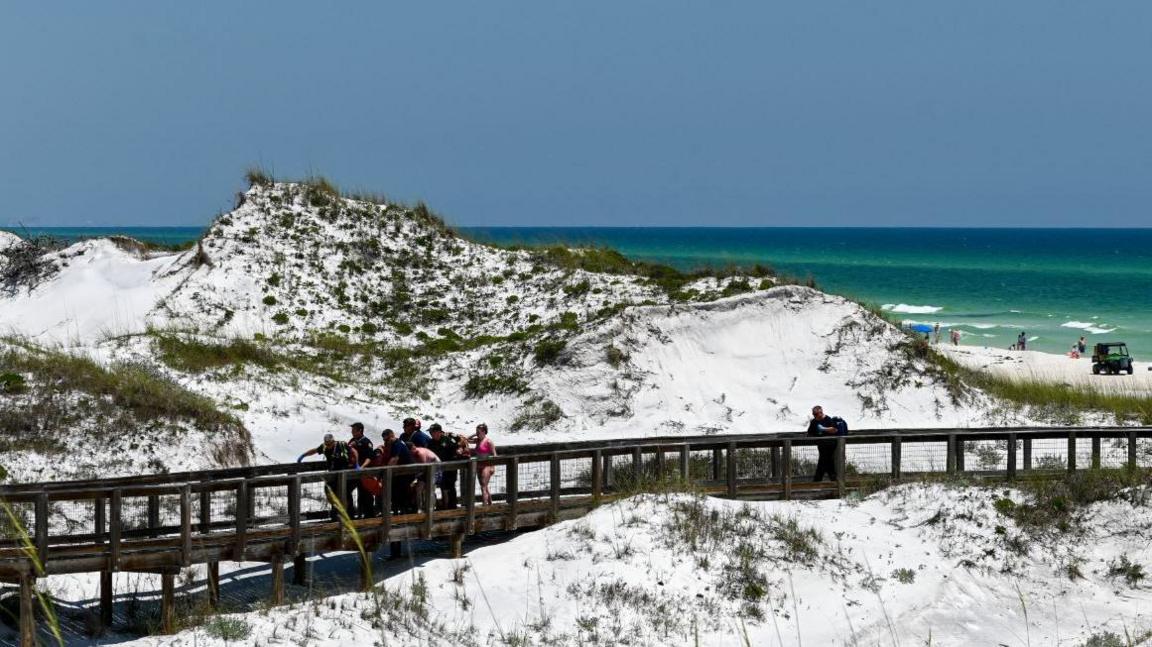 A beach with a walkway, where a crowd of rescuers are surrounding a person being carried on a stretcher