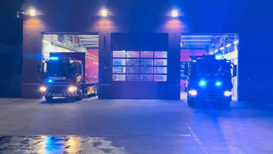 The front of Poole Fire Station at night as two fire engines emerge with their blue lights flashing.