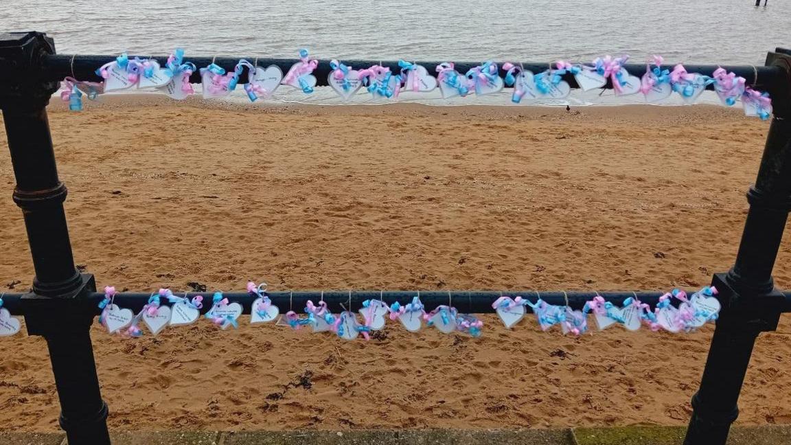 Several white hearts with tributes in writing stuck to a railing on a promenade with blue and pink ribbon and the beach and sea in the background