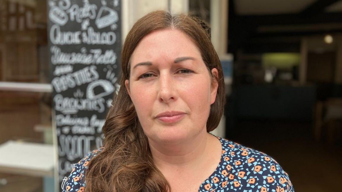 Hannah Lewis in front of the bakery. Her long, brown hair goes past her shoulders and she wears a floral dress