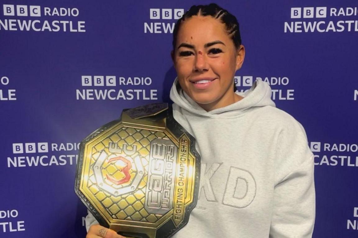 Kennedy Freeman, wearing a cream hoodie and holding her newly won World Championship belt, which is big and coloured gold, silver and steel, with the championship logo on it. She's standing in front of a screen with BBC Radio Newcastle written repeatedly all over it.