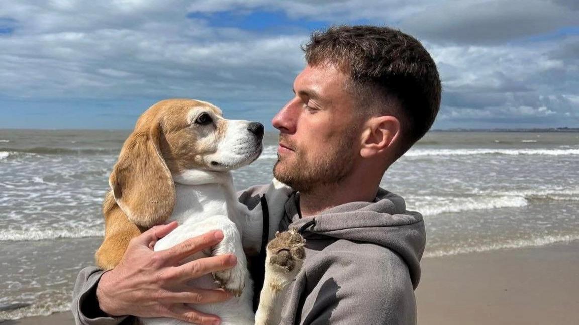 Aaron Ramsey pictured on the beach with sea in background in grey hoodie holding the dog in his arms and looking at it, noses almost touching