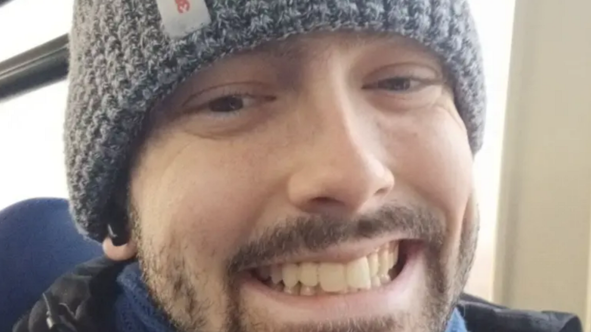 A close-up selfie of a man who appears to be sitting on public transport. He is wearing a grey beanie and smiling at the camera.