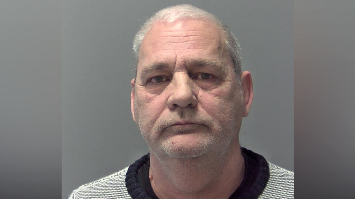 A head and shoulders photo of a man who is looking into the camera. He has short, grey hair and stubble. He is wearing a light grey jumper with a navy neckline. 