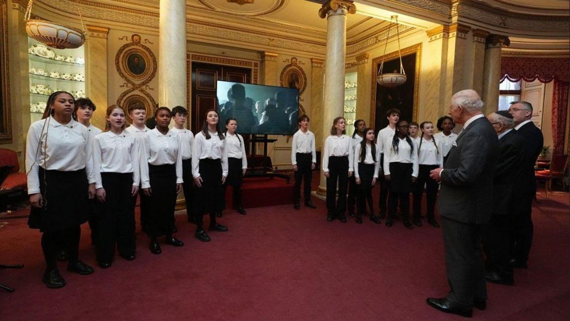 Seventeen students in total are visible on the photo - one group largely to the left of a screen in the middle and another group to the right. King Charles and two other men are on the right of the photo.