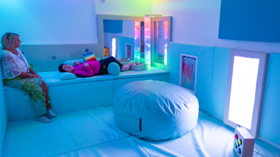 A room filled with soft white furnishings. One person lies on a couch, while a second person sits at the end of it. A large white bean bag is at the forefront of the picture.
