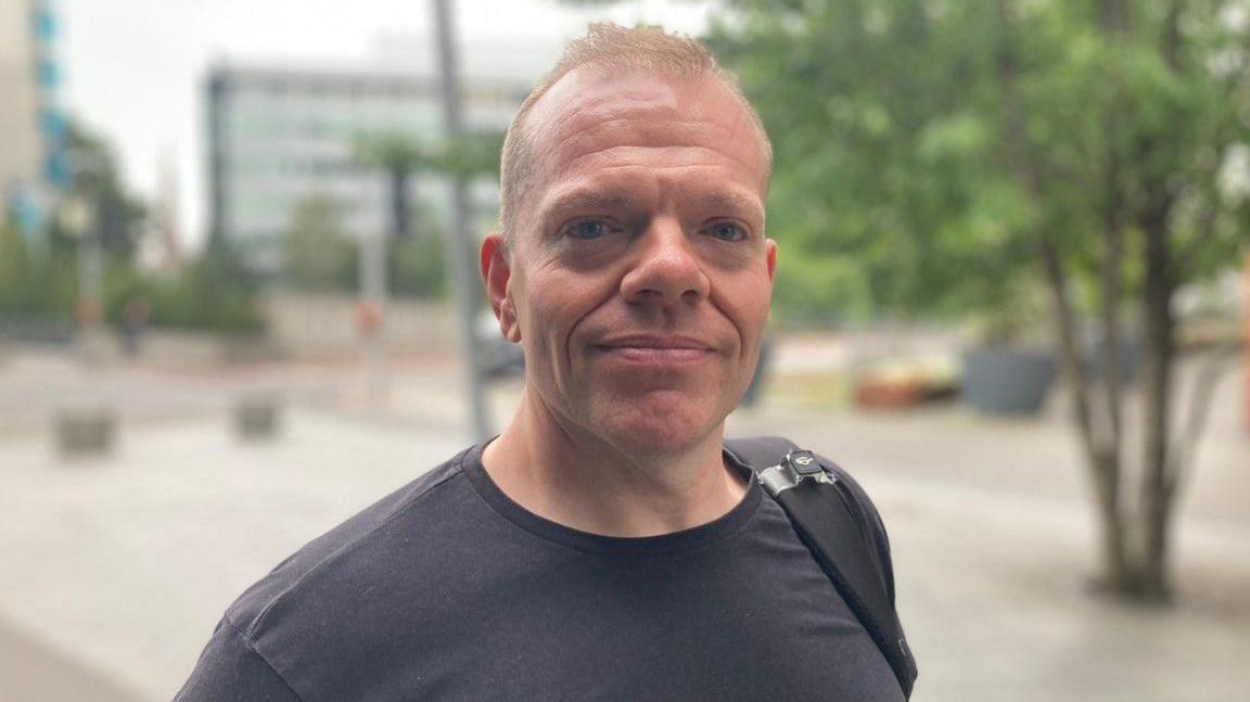A man in a dark top looking at the camera. A tree and buildings are blurred in the background.
