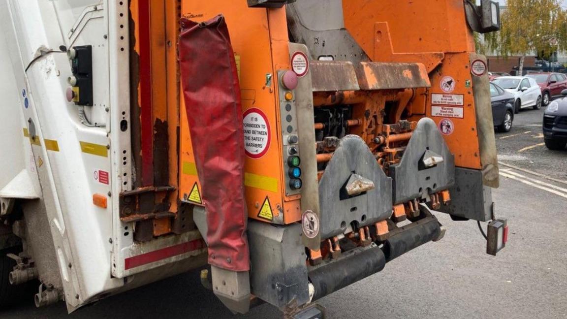 An orange and white truck. It is near cars on a car park.