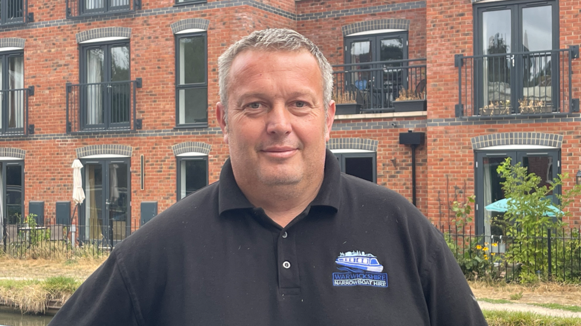 A man with short light brown hair and a black polo top standing in front of a tall brick building with a black metal fence in front of it
