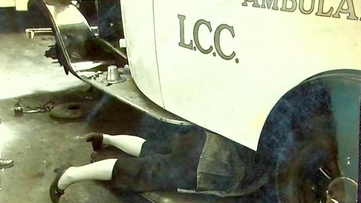 An old ambulance with a woman lying on the floor looking underneath it, with only her waist down visible 