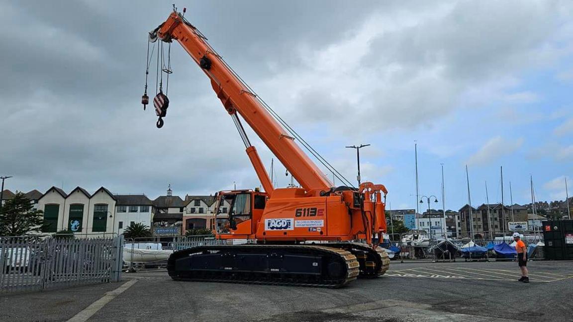 Close up of the crane on a road by railings and a man is stood in high-vis behind it