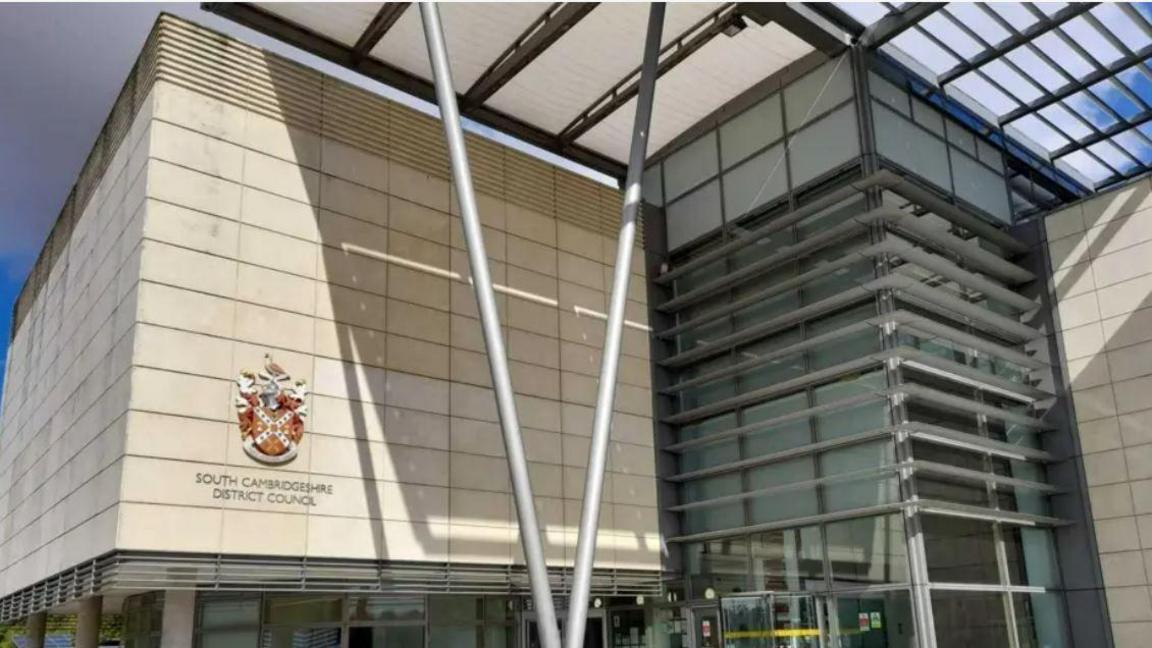 Beige building with a sign saying South Cambridgeshire District Council. two metal bars in a v shape are in front with a glass frontage to the right. 