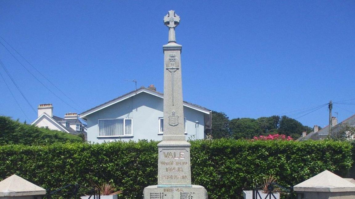 The Vale War Memorial, a granite obelisk with gold and black inscriptions, with a backdrop of a green hedge and a blue sky