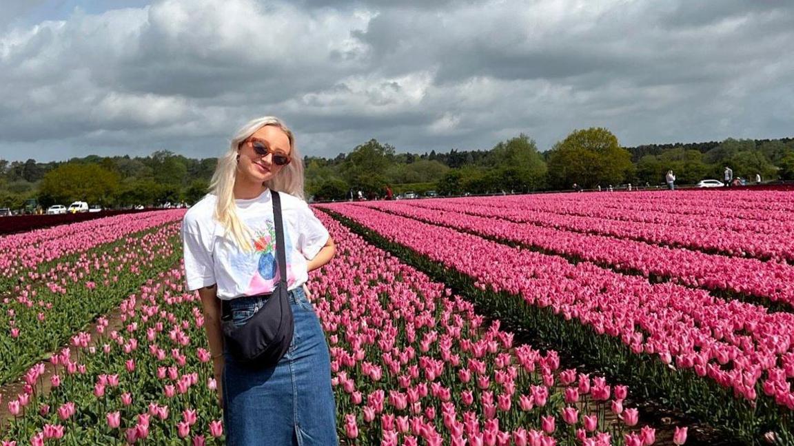 Jamie-Louise Phillips in a white top and blue denim skirt. She has a black crossbody bag on and has long blonde hair and is wearing sunglasses. She is stood in a field of pink tulips.