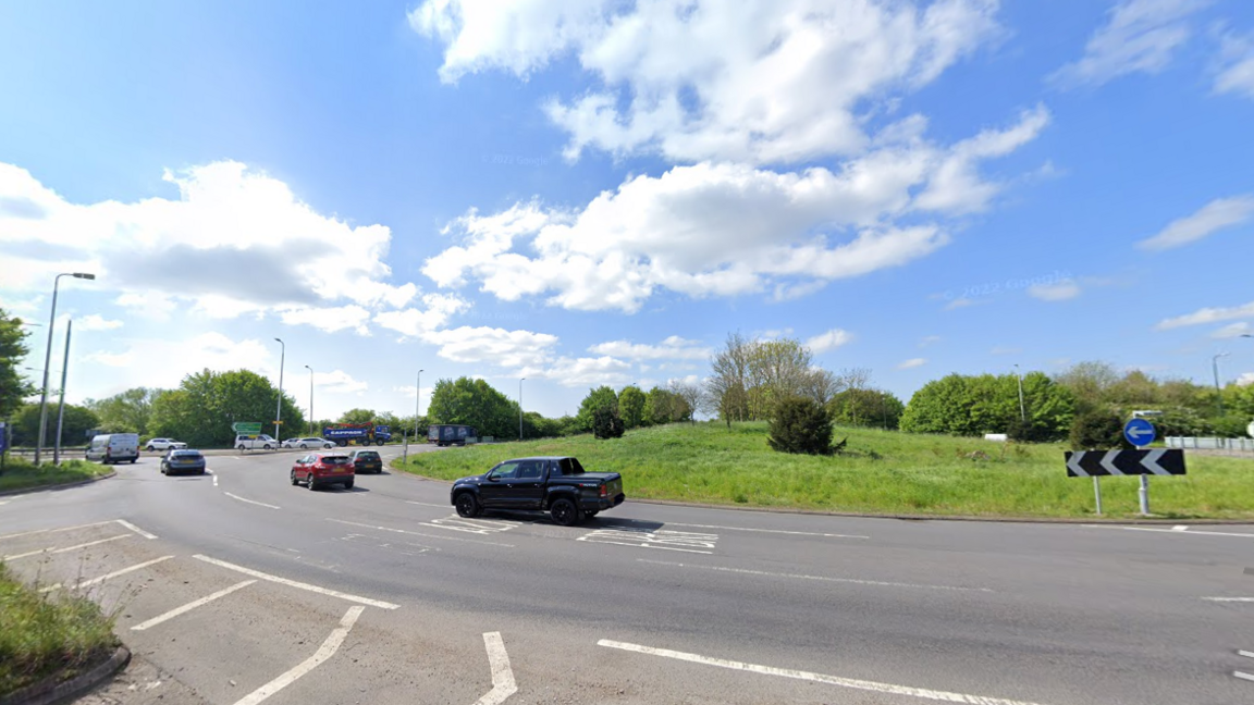 A street view image of a large roundabout with grass in the centre. The sky is blue and cloudy