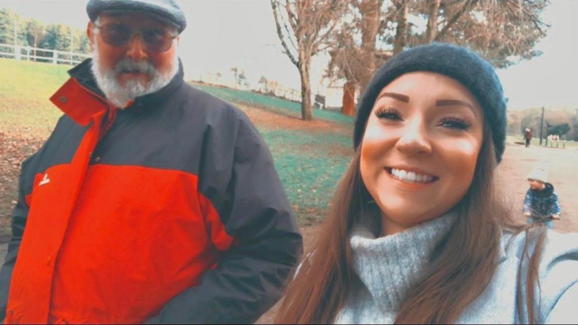 Sian Squires is wearing a bobble hat, walking in the park with her uncle, Stephen who is wearing a flat cap and red rain coat. Both are looking at the camera for a photograph.