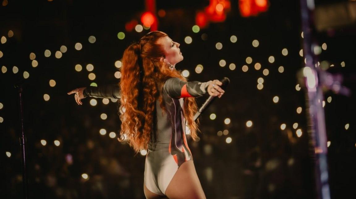 Jess Glynne, with long red hair, performing, with the camera behind her. White lights are in a largely otherwise black background and her arms are outstretched.